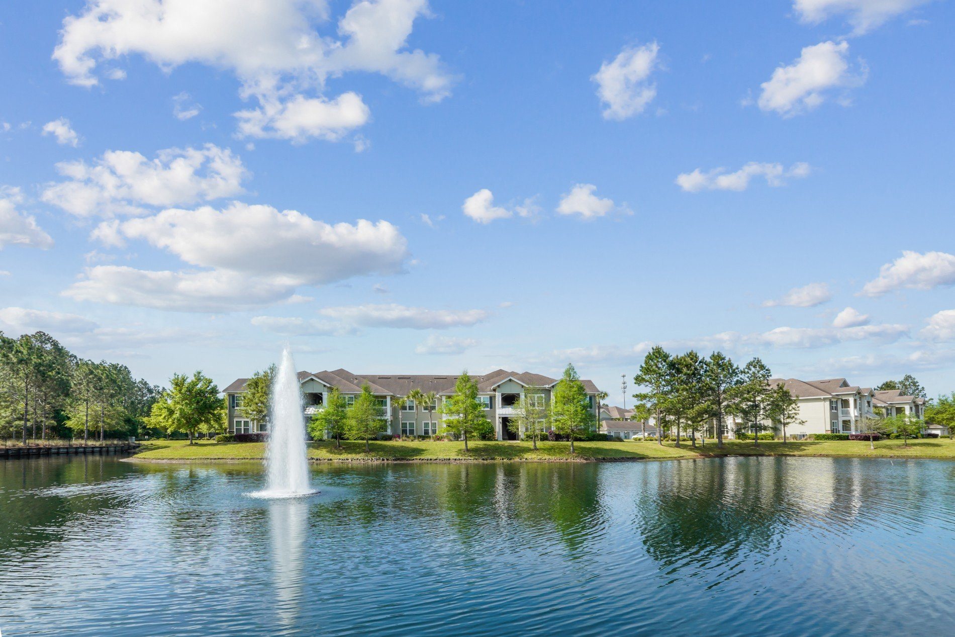 A fountain in the middle of a lake with houses in the background.